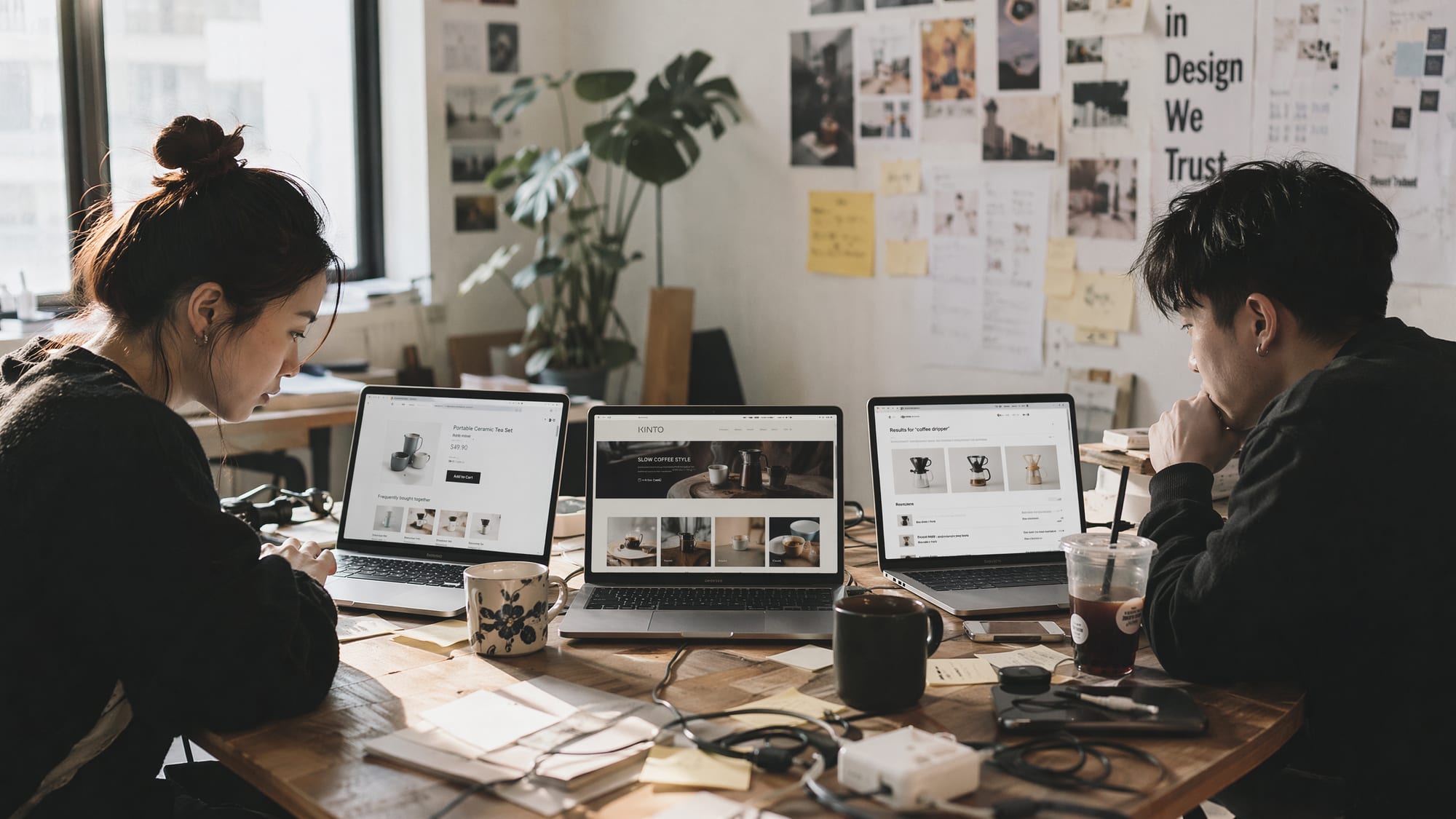 A wide editorial shot of two designers at a daylit studio table, three laptop screens lined up showing an Amazon product detail page with A+ Content, an Amazon Brand Store homepage and a Sponsored Brands banner above Amazon search results, one per service lane.