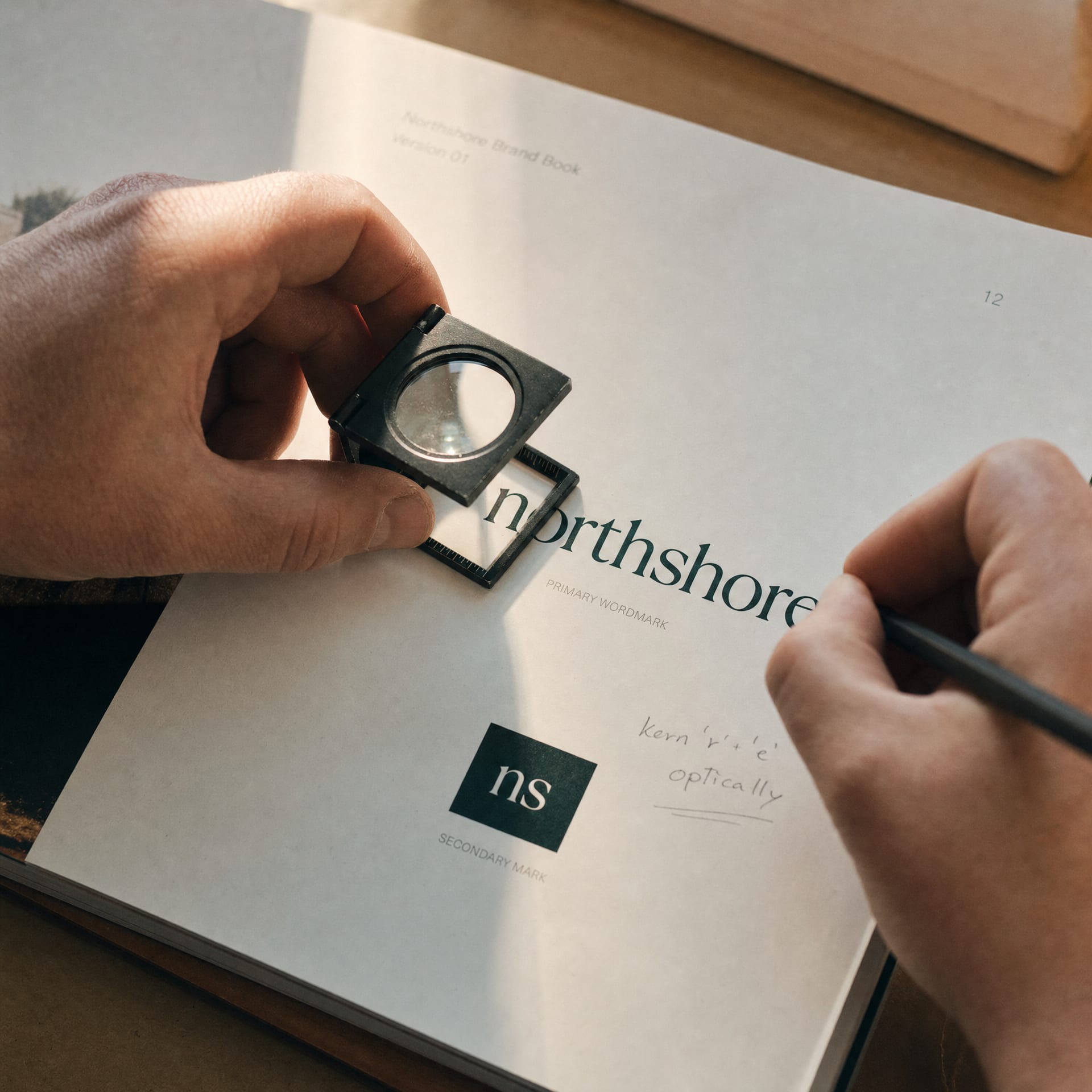 Two hands reviewing a printed brand book proof: one holds a loupe over a wordmark, the other a pencil margin note.