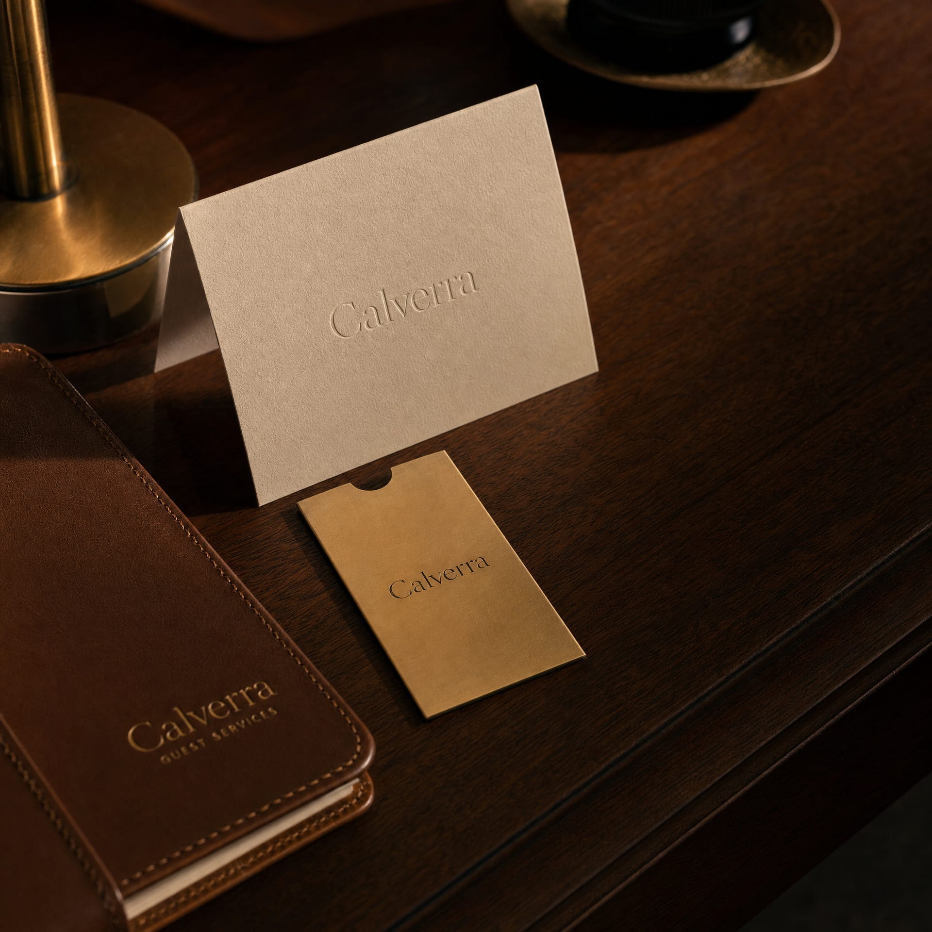 A folded ecru linen card embossed with the Calverra wordmark on a dark walnut hotel desk beside a brass keycard sleeve.