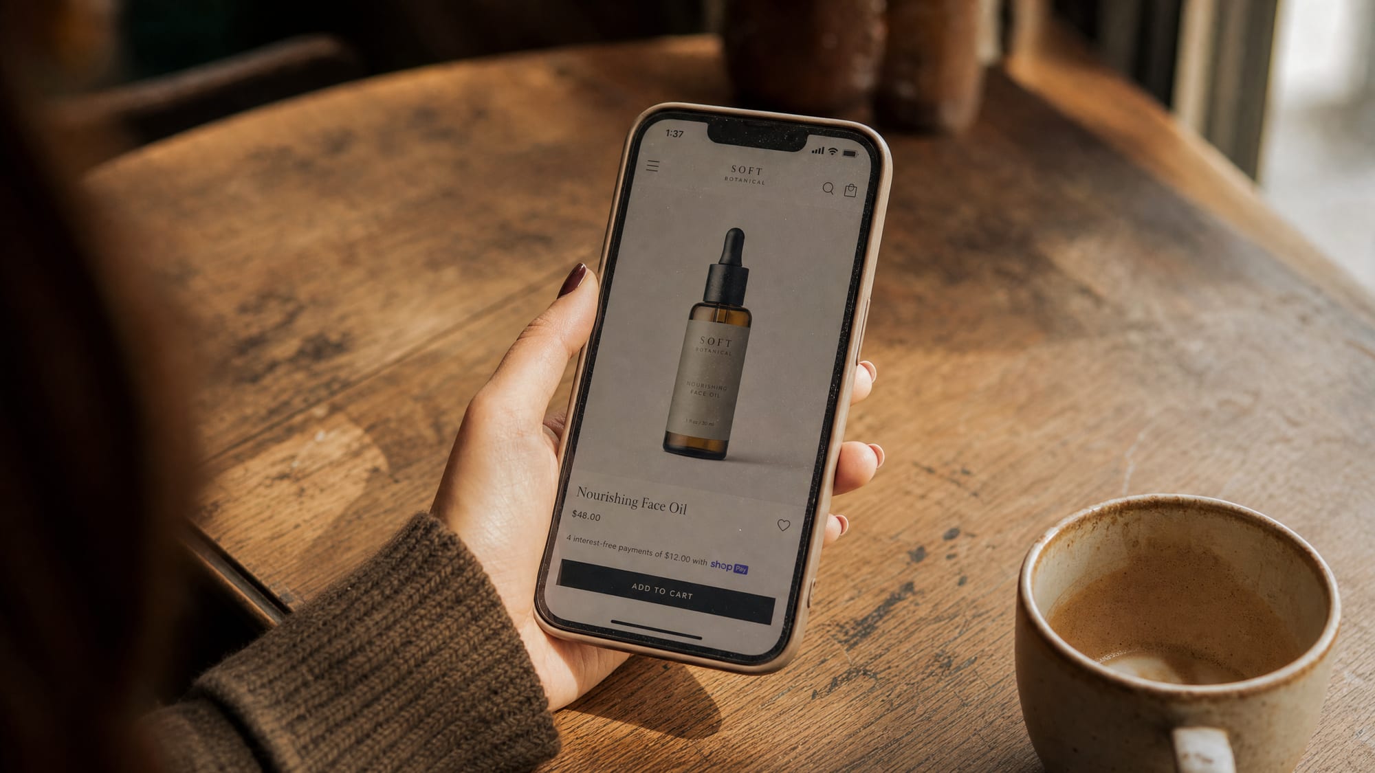 A hand in soft afternoon light holds a phone showing a single beauty product on a vertical mobile shopping listing, on a warm wood cafe table.