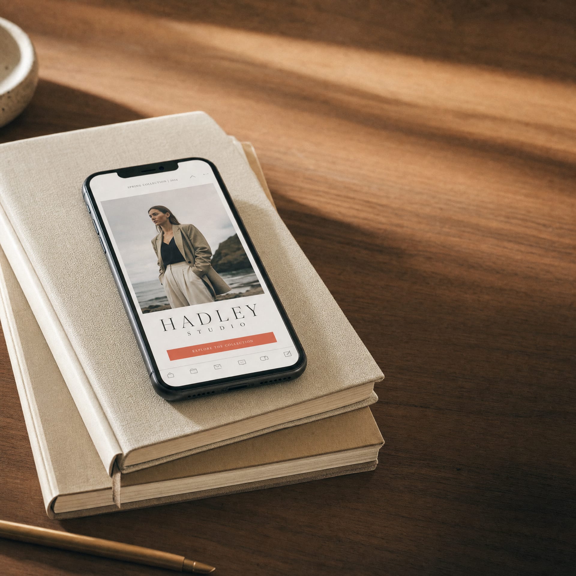A new iPhone on a stack of cream notebooks on a sunlit walnut desk, screen showing an editorial fashion-brand newsletter email.