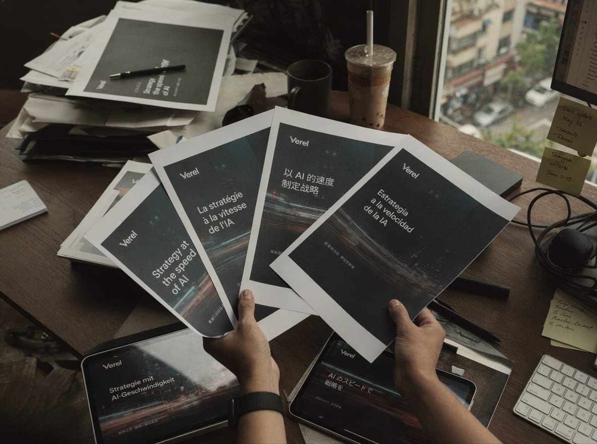 Four printed deck proof sheets and two tablets arranged in a soft fan on a warm walnut desk, each showing the same slide in a different language and aspect ratio.