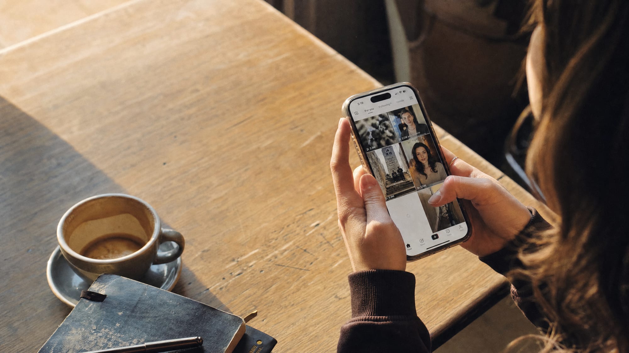 Hands holding a vertical smartphone scrolling a social feed at a sunlit cafe table.