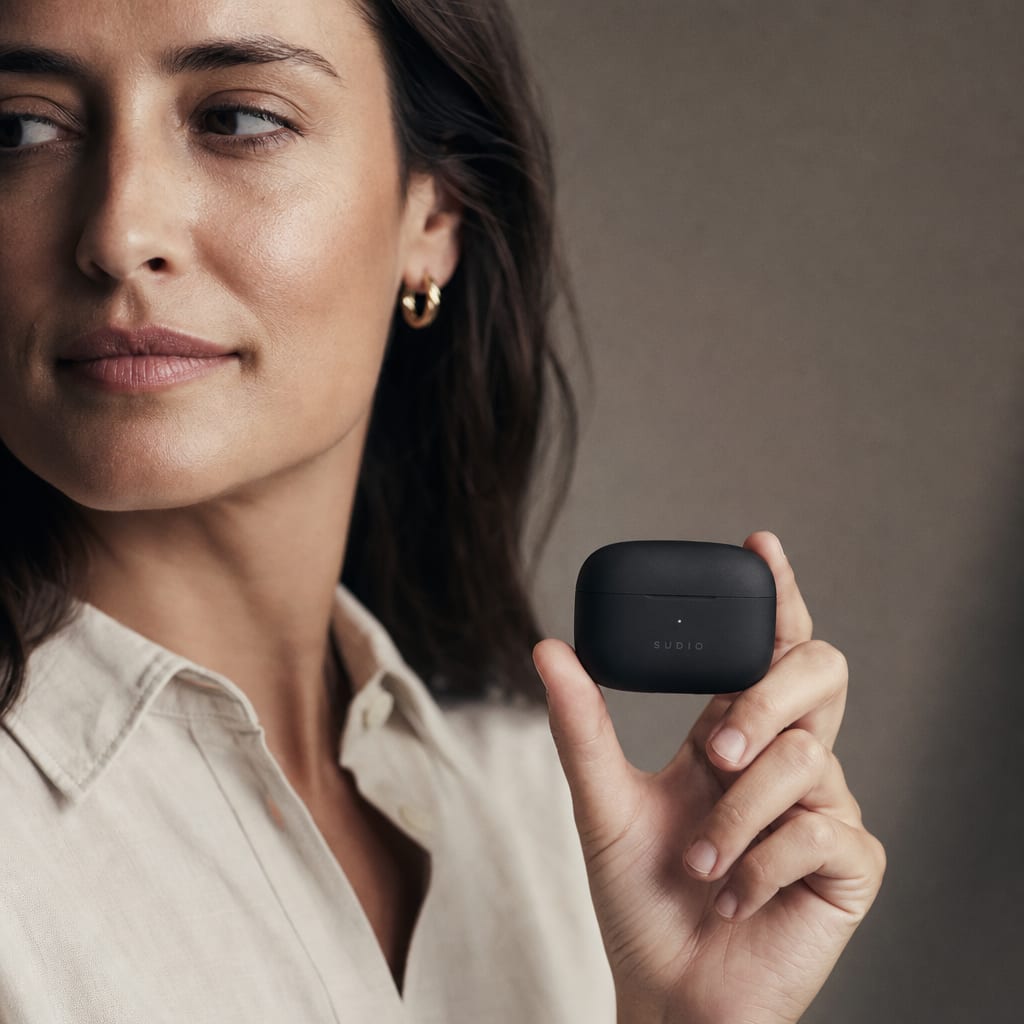 A woman holding a matte black wireless earbud case at eye level under one directional studio light.