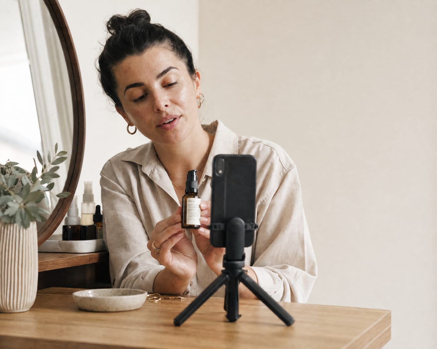 A beauty creator at a vanity desk holding a skincare serum bottle to a vertical phone on a tripod, filming a how-to in soft window light.