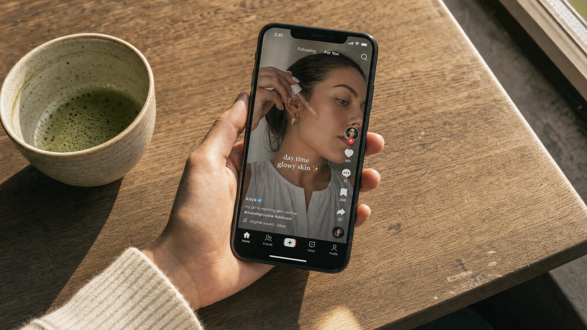 A hand holding a vertical phone showing a TikTok-style feed in mid-afternoon café light.