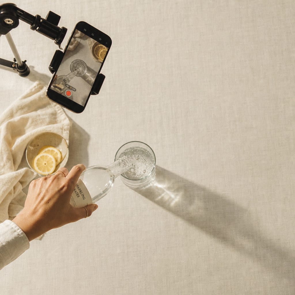 Overhead shot of a creator pouring sparkling water with a phone on a clamp filming the pour.
